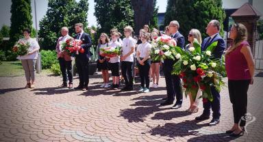 Na zdjęciu widać kilkanaście osób, niektóre z nich trzymają kwiaty